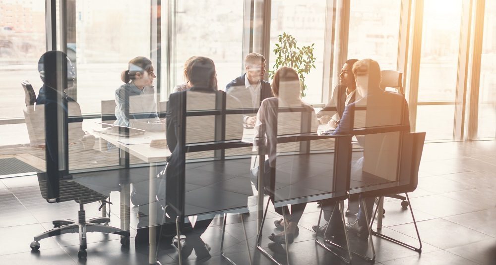 8 Menschen sitzen hinter einer Glaswand an einem Konferenztisch