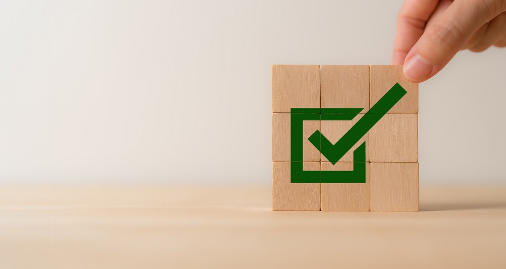 9 Holzklötzchen aufgestellt als Quadrat mit einem Kästen drauf mit Häkchen und eine Hand oben am Quadrat