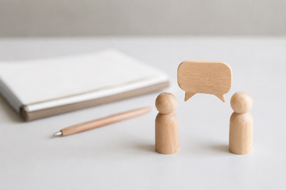 Zwei Holzfiguren mit einer Sprechblase, Block und Stift im Hintergrund
